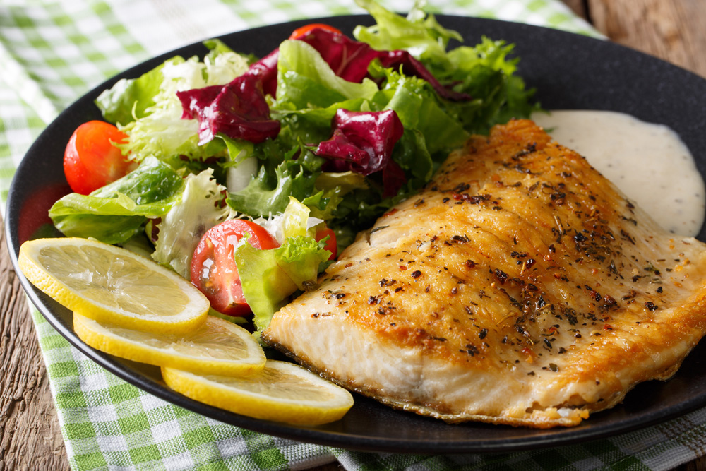 Close-up photo of a fillet of salmon garnished with lemon and a side green salad served on a black ceramic plate