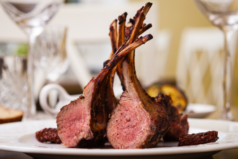 Photo de deux carrés d’agneau mi saignants servis dans un plat, et de verres à vin en arrière-plan